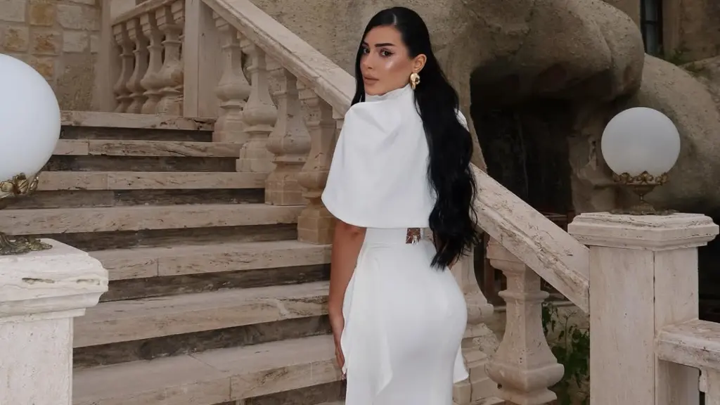 Kader Kicaj e veshur me fustan të bardhë elegant duke pozuar në shkallë prej guri, me një atmosferë elegante dhe përrallore.