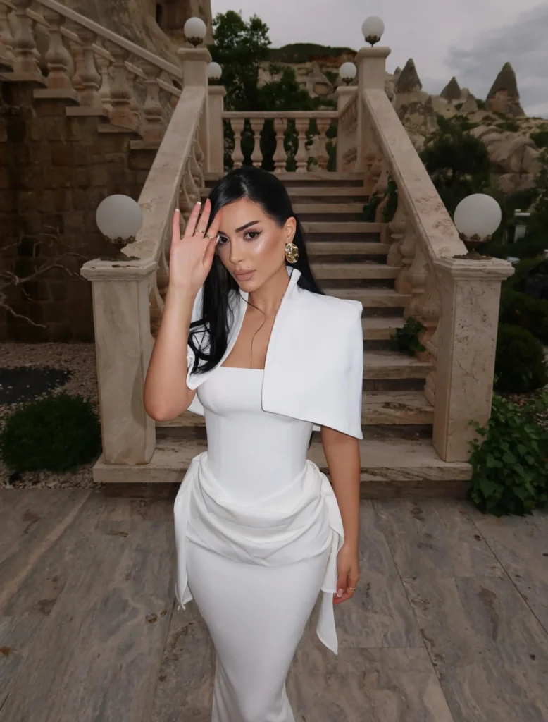 Kader Kicaj e veshur me fustan të bardhë elegant duke pozuar në shkallë prej guri, me një atmosferë elegante dhe përrallore.